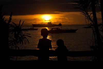 Silhouette boy sitting on beach against sky during sunset