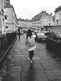 Full length rear view of woman with backpack walking on wet sidewalk amidst buildings