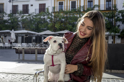 Young woman smiling while sitting with dog on chair in city