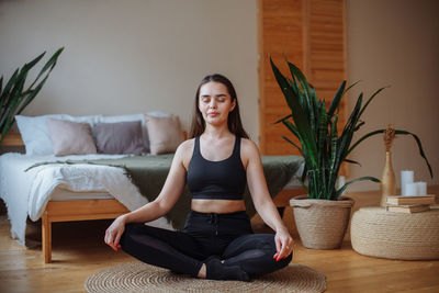 A young woman sits at home on a jute rug near the bed in the lotus position. does yoga at home