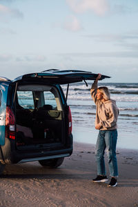 Young woman standing by car at beach against sky during sunset