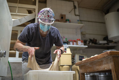 Unrecognizable female baker in mask working in bakery and kneading raw dough while cooking delicious bread