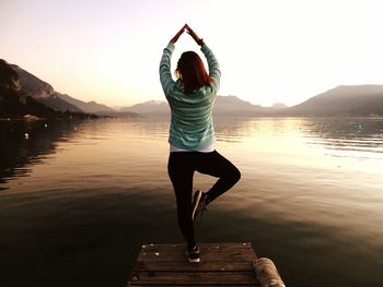 Rear view of woman doing yoga on pier over lake against sky during sunrise