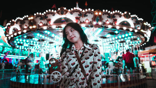 Portrait of young woman standing in illuminated carousel at amusement park