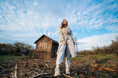 Beautiful girl with long hair in a grey trench coat next to an old wooden house blue sky background 