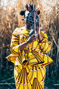 African woman in yellow dress with authentic print covering face with horse mask while standing near dry grass in field
