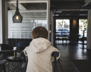 Rear view of boy wearing hooded jacket while leaning on chair in cafe