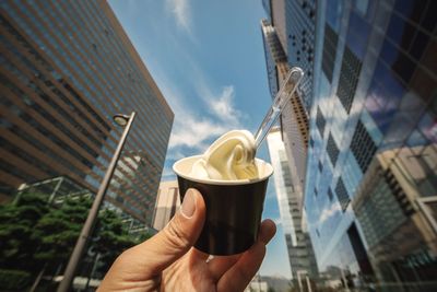 Cropped image of hand holding ice cream cup against buildings