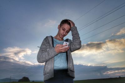 Young man using smart phone while standing against cloudy sky