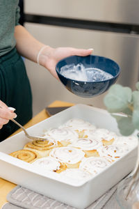 A young girl spreads cream on the cinnabons in a baking dish on the table.