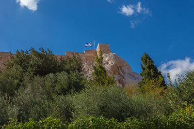 Acropolis walls of athens viewed from below with trees and greek flag flying, greece