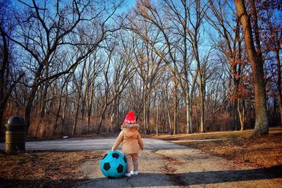 Rear view of boy standing by soccer ball on road against bare trees during winter