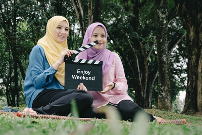 Low angle view of happy friends holding film slate while sitting against trees in forest