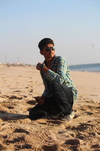 Young man sitting on sand at beach against clear sky