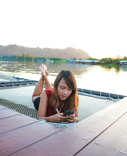 Woman using phone while lying on net by lake