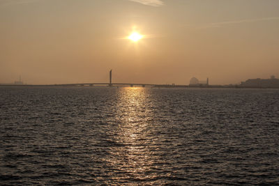 Distant view of silhouette bridge over sea against orange sky