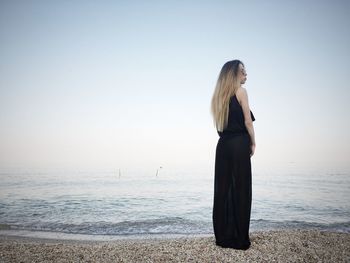 Full length of young woman wearing black dress while standing on shore at beach against sky