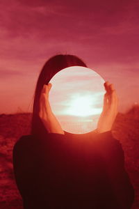 Woman holding mirror with reflection while standing against sky during sunset