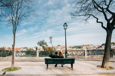 Rear view of couple sitting on bench against sky