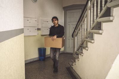 Full length of delivery man with package box walking by staircase