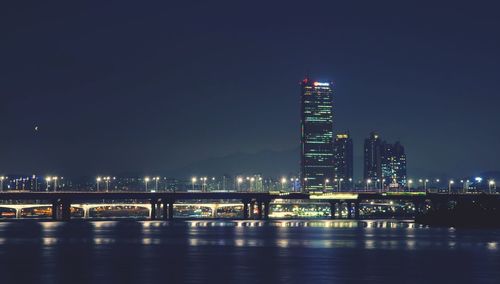 Illuminated bridge over river by buildings against sky at night