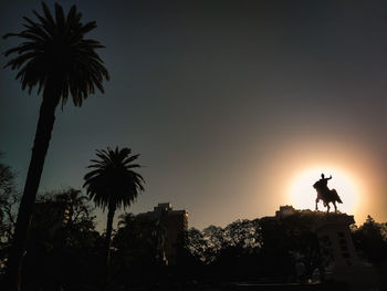 Silhouette statue by palm trees against sky during sunset