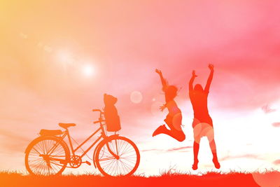 Silhouette siblings with arms raised jumping by bicycle on field during sunset