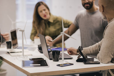 Business people discussing over windmill while using technologies at desk in open office space