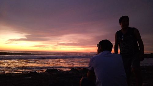 Rear view of friends standing at beach against sky during sunset