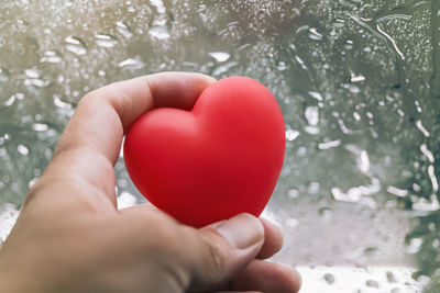 Cropped hand holding red heart shape by wet window