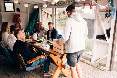 Young man holding crate while enjoying lunch party with friends in cottage