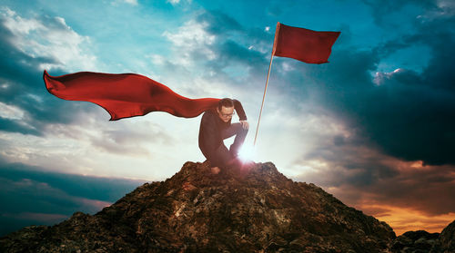 Low angle view of man in superhero costume kneeling by red flag on rock against sky