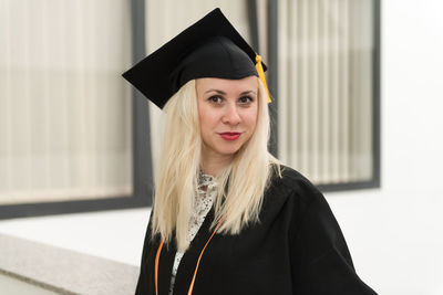 Young woman wearing graduation gown standing against wall