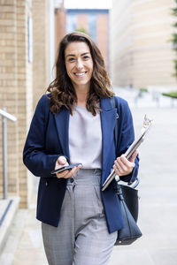 Smiling businesswoman with laptop and smart phone walking on footpath