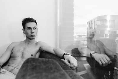Shirtless thoughtful man siting on sofa by window at home