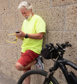Bearded senior man using mobile phone while standing by bicycle against wall
