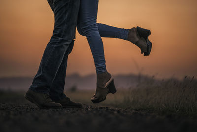 Low section of couple standing on land against sky during sunset