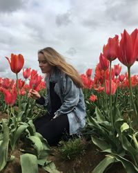 Young woman looking flowers while crouching on field against sky