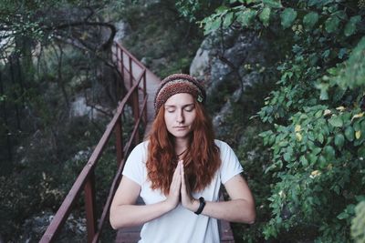 Beautiful young woman with hands clasped on footbridge against trees