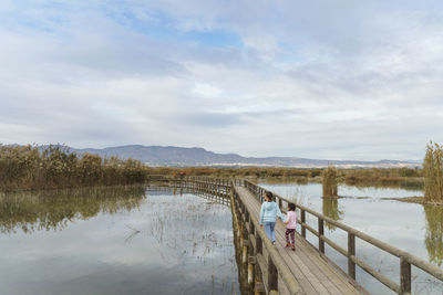 Pier over lake against sky and two girls running on a bridge 