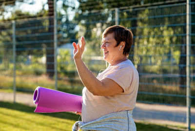 Happy smiling senior plus size woman with earphones holding yoga mat outdoors waving bye to coach 