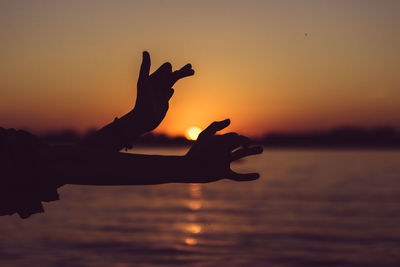 Cropped hands of silhouette person gesturing over sea against sky during sunset