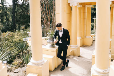 A beautiful young man, the groom in an elegant wedding suit, stands posing in the city's old park