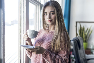 Portrait of smiling young woman standing against window at home