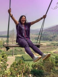 Full length portrait of woman sitting on swing against sky