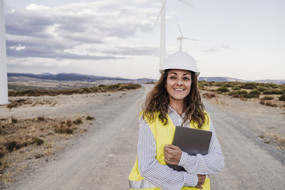 Smiling engineer wearing hardhat holding laptop at wind farm