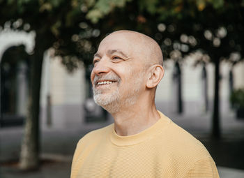 Elderly man in yellow sweater smiling contentedly in urban tree-lined avenue