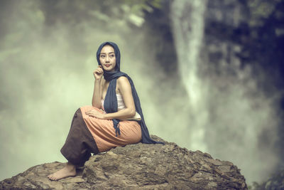 Portrait of young woman sitting on rock against trees