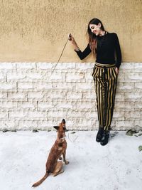 Happy young woman showing stick to puppy while leaning on wall