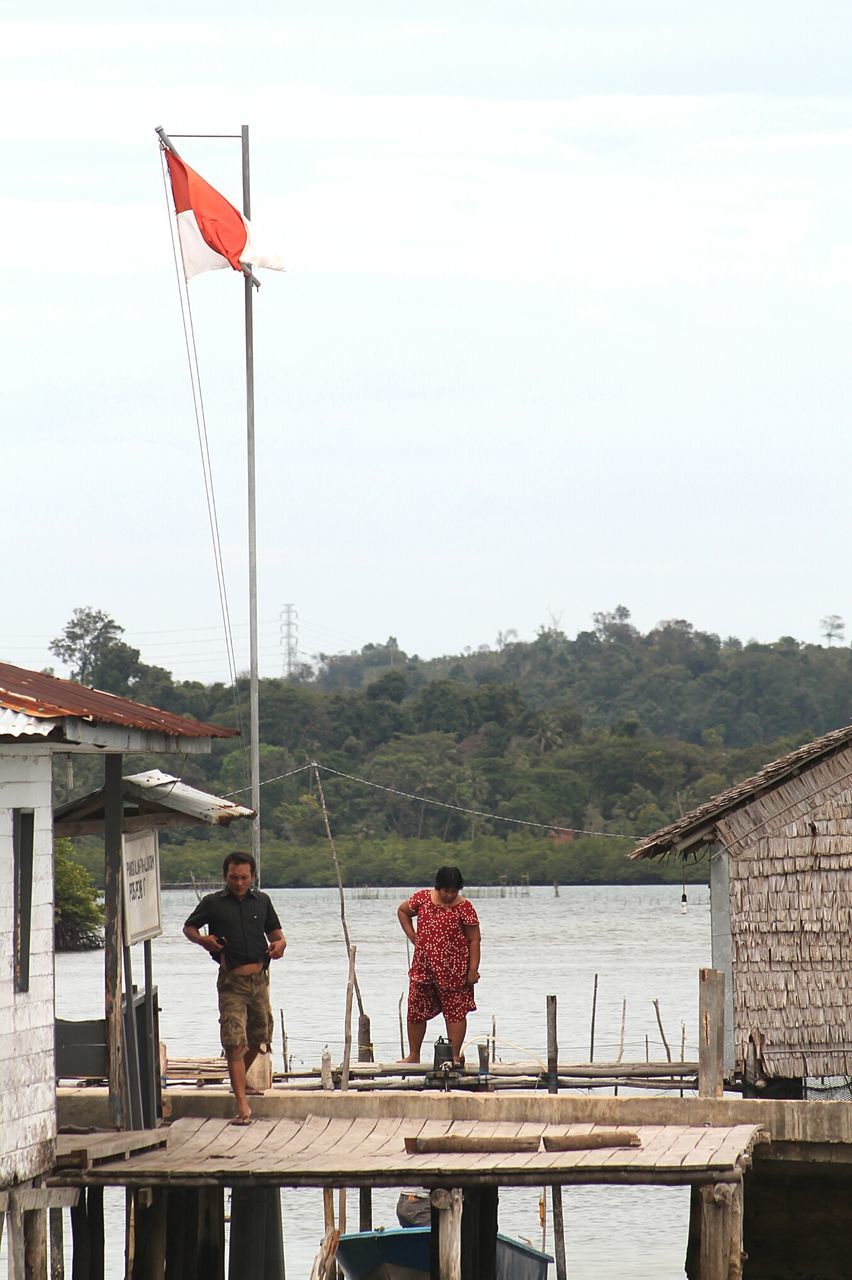 lifestyles, leisure activity, rear view, railing, mountain, flag, men, full length, sky, red, water, person, national flag, standing, casual clothing, day, built structure, patriotism